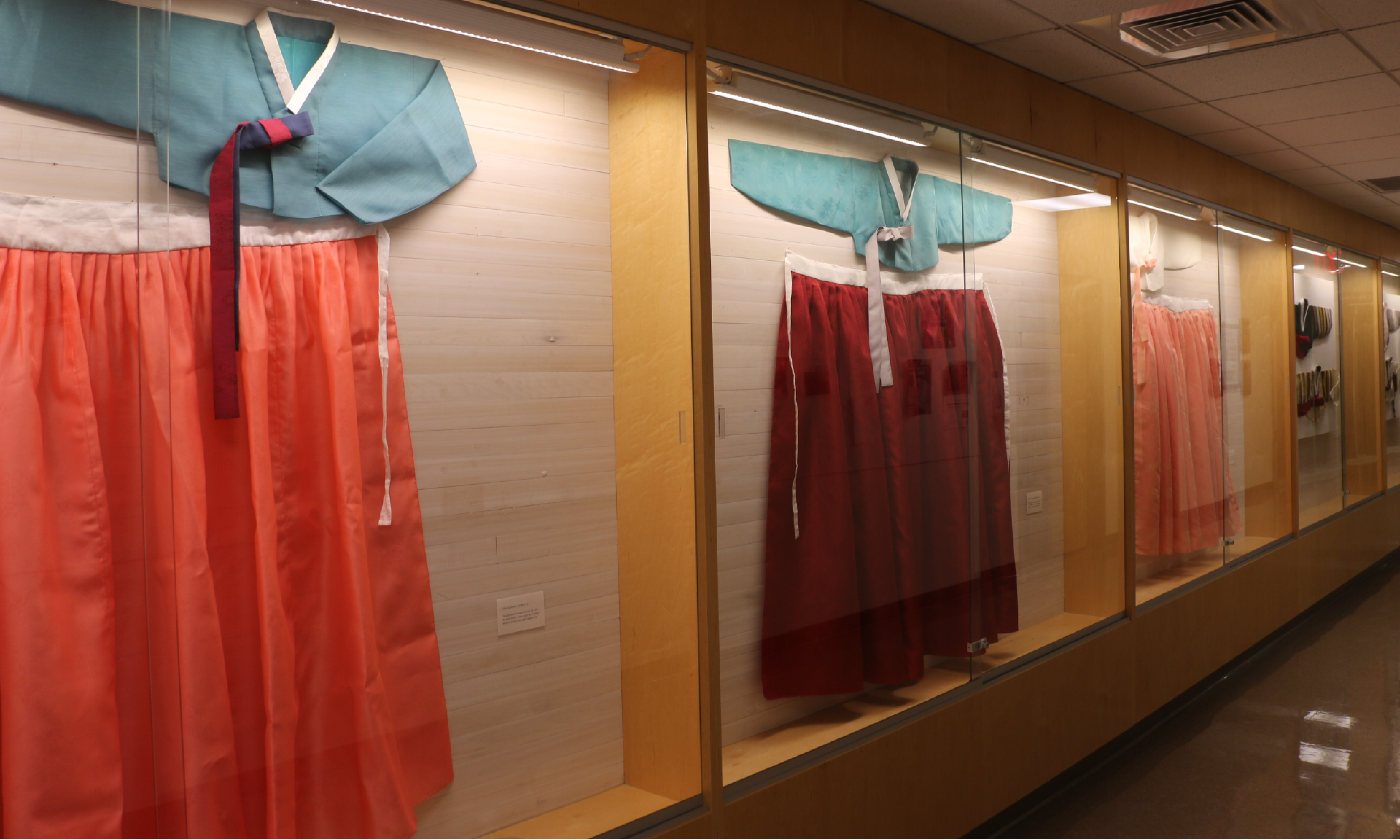 Three blue, red, and pink hanbok hang inside of glass cases in a hallway.