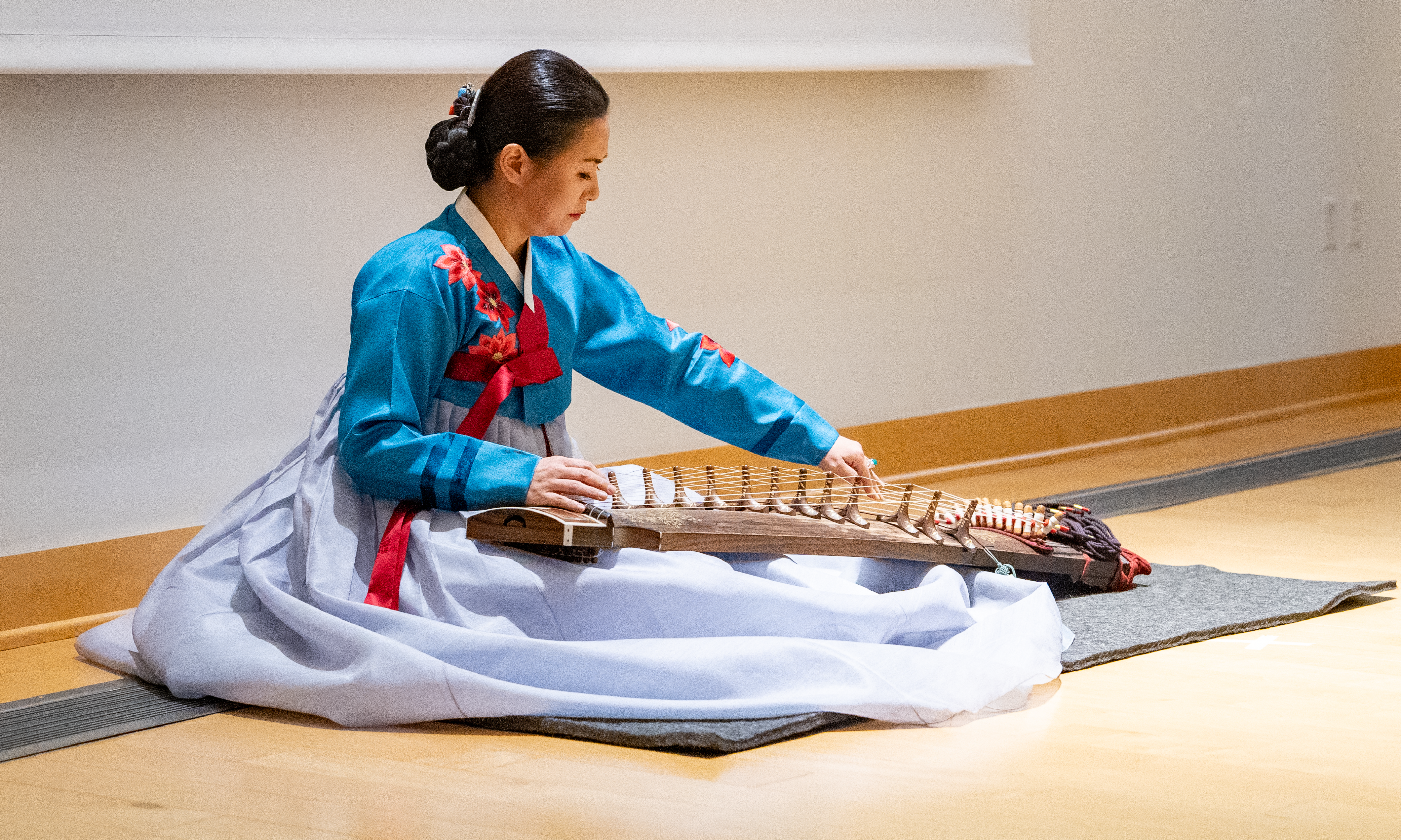 A woman sits on a mat on the floor and plays a gayageum, a string instrument. 