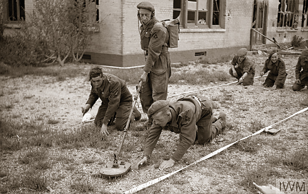 An old, black and white photo of several soldiers from World War II using a mine detector. Several soldiers are kneeling while one stands and operates the detector. 