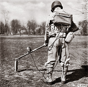 An old photo of a World War II soldier from behind with a metal detector.