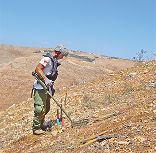 A man in personal protective gear holds a metal detector on a rocky hillside.