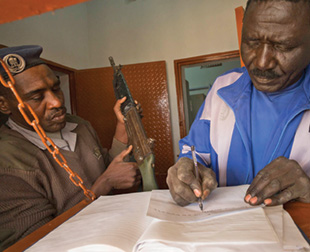 A man wearing fatigues and a beret holds a rusty rifle next to a man in a blue jacket who writes on a piece of paper.
