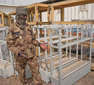 A man in fatigues and a scarf gestures by white storage racks