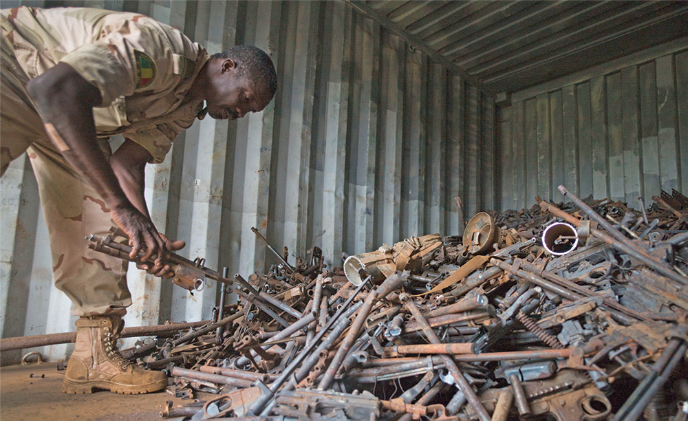 A man in fatigues leans over and collects rusty weapon components.