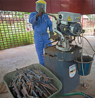 A man in a blue jump suit and yellow headwear operates a machine by a container of weapon components.