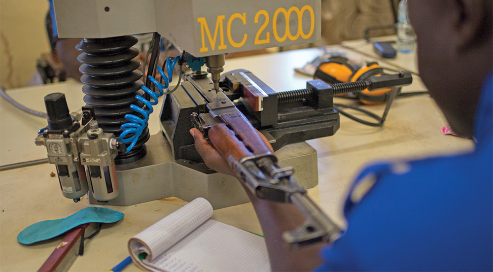 A rifle in a machine with the label "MC 2000" in front of a man with a blue shirt.