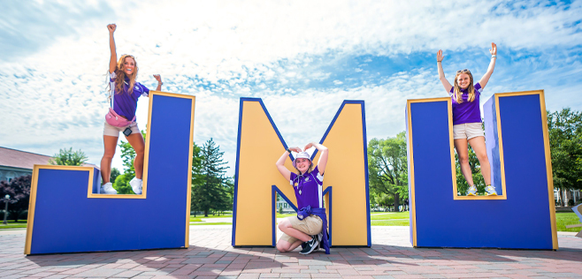 Three girls stand on top of oversized JMU letters