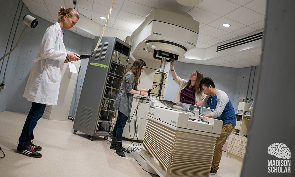 Four people in the lab, one standing off to the left of the acclerator machine wearing a white lab coat and holding a clipboard; and the other three wokring around the large machine.
