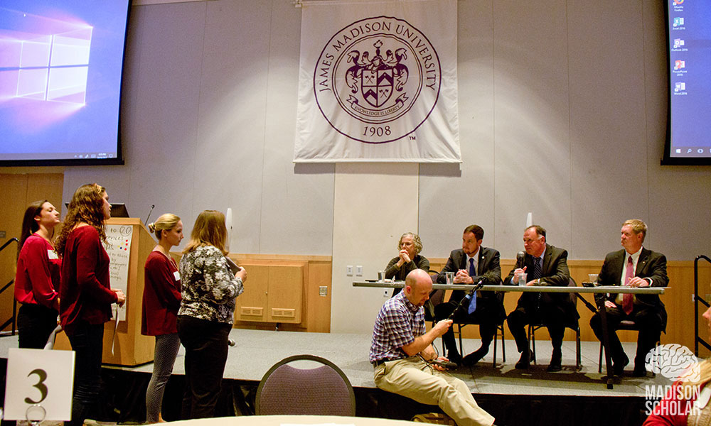 Panelists behind a table on a stage listen as students stand and discuss their proposal in front of the stage.