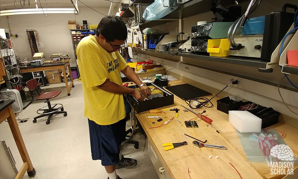 Premal Patel, dressed in yellow t-shirt and dark shorts, works on a power box at a work bench.