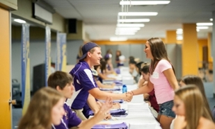 male student in purple on one side of long registration table shaking hands with female student who is on the other side