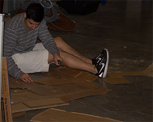 student sitting on the floor, looking down at project, surrounded by pieces of cardboard.