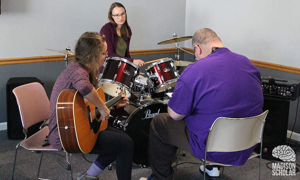 a student on drums and another with a guitar work with a Gemeinschaft resident who is also seated with his back to the camera.