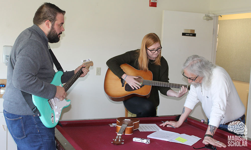 a professor and a student with guitars work with a Gemeinschaft resident around a red pool table.