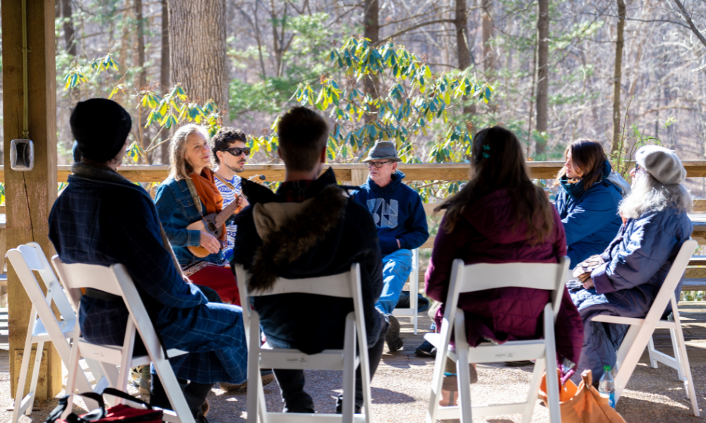 Nine people in coats sit in white chairs in a circle outside at the Arboretum Pavilion. A blonde woman holds a ukulele and leads the group in song. 