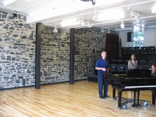 A grand piano sits on the hardwood floor in the performance area with the bluestone wall in the background.
