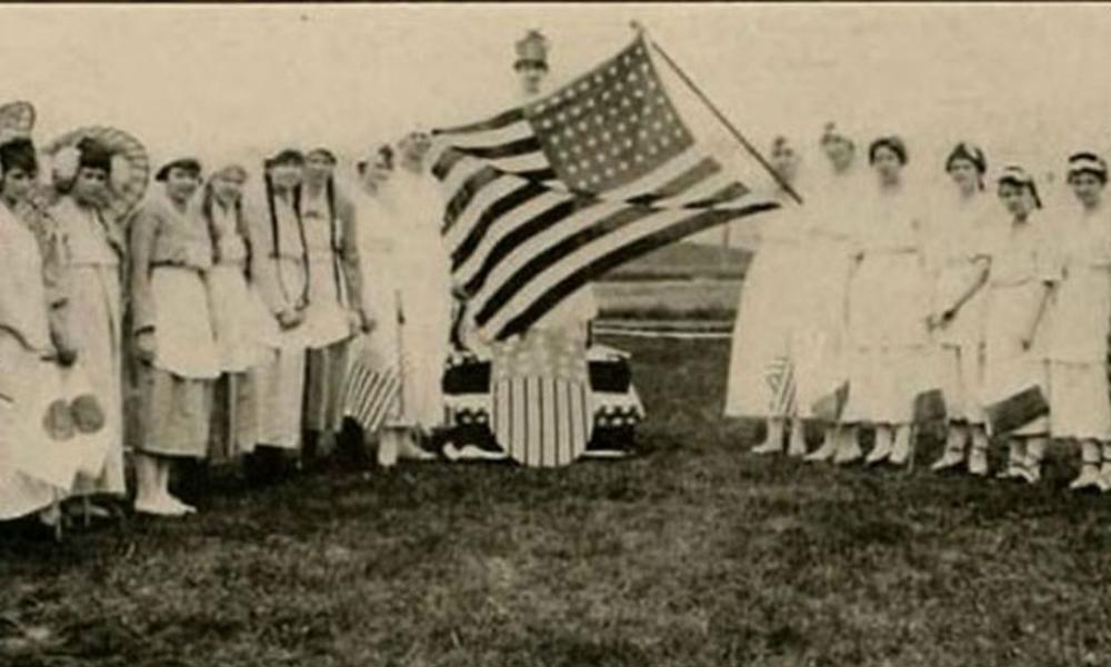 an old sepia-tone photo of ladies of at JMU posing around a US flag with 48 stars (pre 1959)