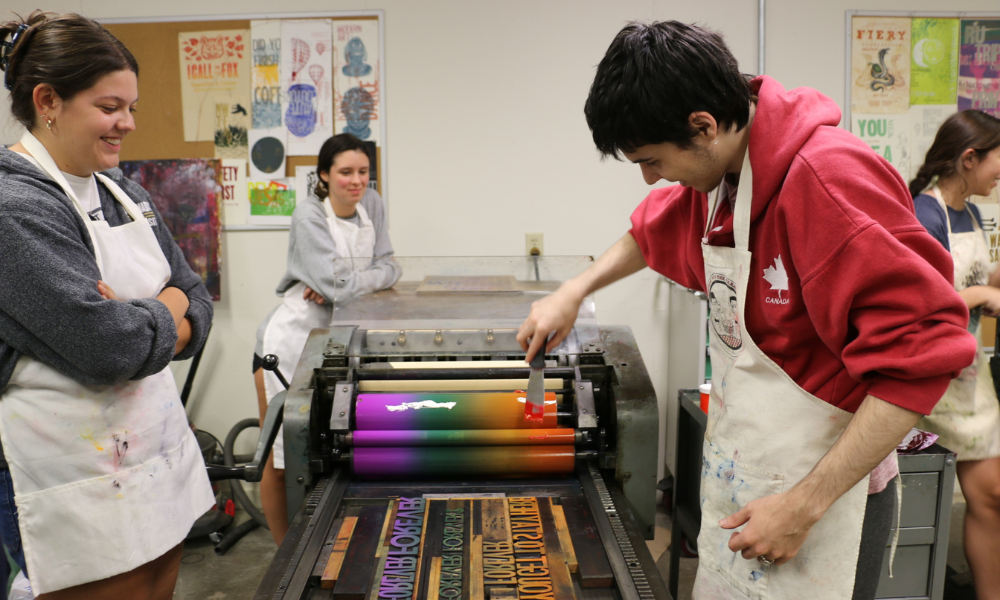 Three students clean ink off of the roller on the printing press.
