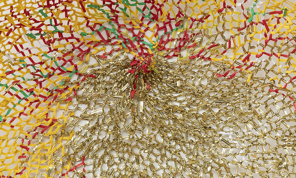 Close-up image of a flat circular structure composed of gold, red, yellow, and green twist ties. 