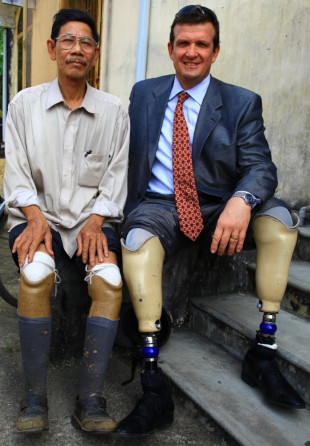 Ken Rutherford (right) sits with another man (left) on the edge of dark steps. Both men's pant cuffs are rolled up to the thigh to show their prosthetic legs.