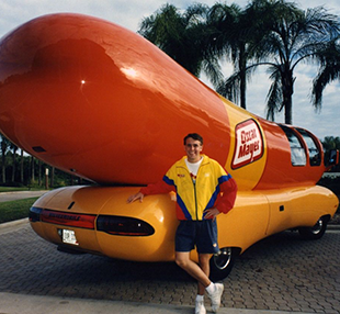 A young man in a colorful windbreaker leans against the Oscar Mayer Weinermobile.