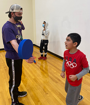 A young man holds out a punching target for a younger child in a gymnasium.