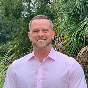 A portrait of Justiss Wilder Burry, with very short hair and a pink button-up, in front of a green, leafy forest background.