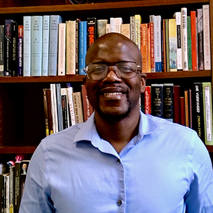 A portrait of Akil A. Cornelius Jr., wearing a blue button up standing in front of packed bookshelves.
