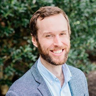 A portrait of Ian Hensley, smiling widely in front of a leafy green background.