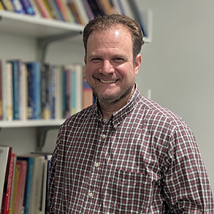 A portrait of Jeremy Rosselot-Merritt, with short brown hair in a checkered button-up, standing in front of a wall of full bookshelves.