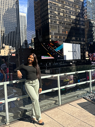 Sheridan Osei leaning against a railing of a glass-walled building in New York City.