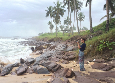 A young woman stands alone on a rocky beach, pointing a camera at the ocean as she takes a photo.