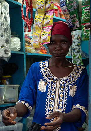 An African woman manning a storefront, products brush her forehead as she hands over change to a customer.