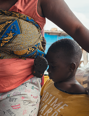 A child leans his forehead against a woman's hip. The woman is carrying another young child in a wrap around her chest. 
