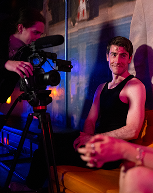 A young man leans against a wall speaking to someone out of frame while another person adjust a film camera pointed at him.