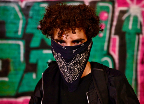 A young man stares into the camera. A black bandana covers the lower half of his face. The wall behind him is covered in graffiti. 