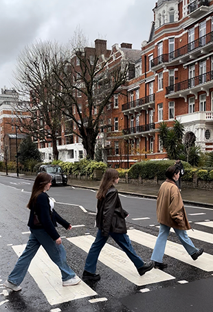 Three girls walk in a line, recreating the iconic Abbey Road crosswalk photo from the Beatles album cover.