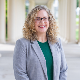 A woman with shoulder-length curly blonde hair wearing a gray blazer over a green blouse.