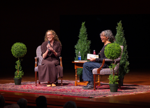Two women sit in chairs on a stage decorated with greenery.