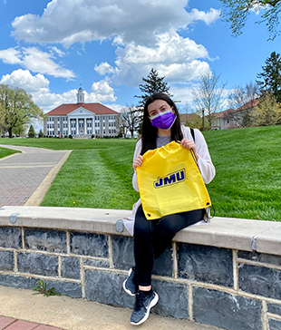 A young woman wearing a purple facemask holds up a yellow bag with a purple JMU logo, sitting at the bottom of the quad with Wilson Hall visible in the distance.