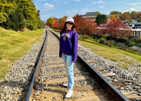 A young woman in a JMU sweatshirt and ballcap stands on railroad tracks.