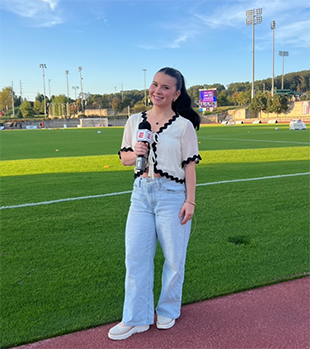 A young woman holding a microphone stands in front of an empty soccer field.