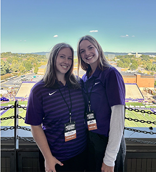 Two people with press box badges stand next to a short wall, with Bridgeforth Stadium stretching out below.