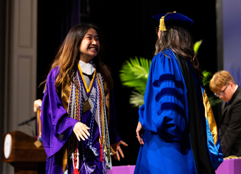 A young woman in purple graduation regalia smiles widely as she shakes the hand of a person in doctorate regalia.