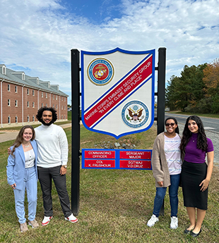 Four people stand next to a sign, two on either side, that reads Marine Corps Embassy Security Group "In every clime and place'