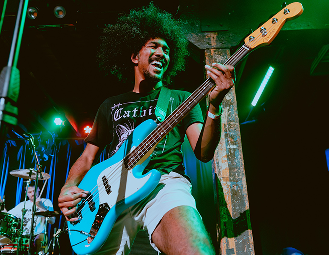 CJ Uy, a Black man with an afro, plays a blue electric guitar, smiling broadly.