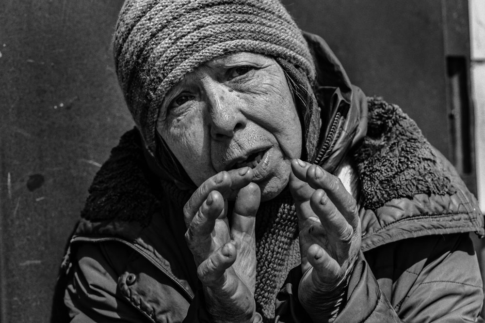 An older woman in a coat and hat with her hands up near her face,