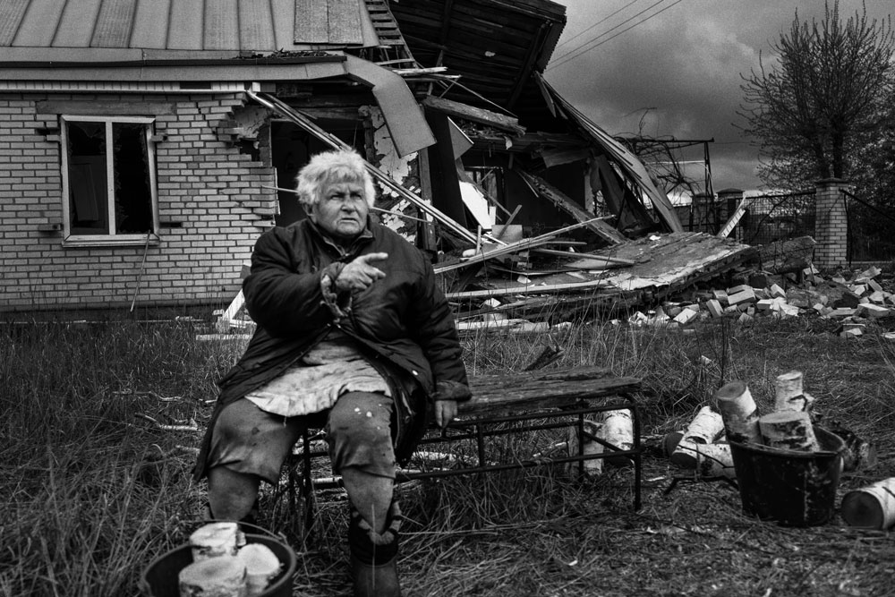 An older woman sits in a chair in front of her damaged house.