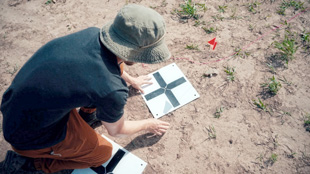 Setting up a ground control point for unmanned aerial vehicle (UAV)-based controlled experiments at Oklahoma State University’s Center for Fire and Explosives (CENFEX). Note inert PFM-1 in the background.  Image courtesy of Jasper Baur.