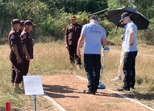 A group of men standing around someone holding a metal detector and walking down a dirt lane.